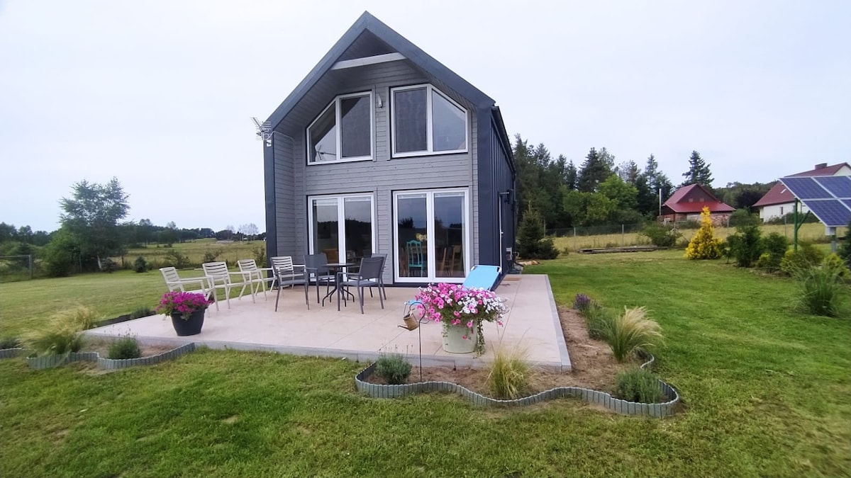 The exterior of the house is presented, featuring a two-story design with large windows. A spacious patio area is surrounded by flower beds containing various plants. Outdoor seating is arranged for relaxation, complemented by a grassy area that enhances the setting.