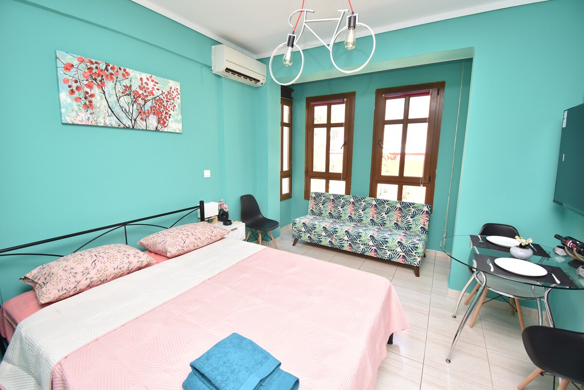 A bright bedroom features soft turquoise walls and large windows allowing natural light. A spacious bed is adorned with pastel-colored pillows, while a patterned sofa and dining table complement the space. A wall art piece adds a touch of color above the bed.