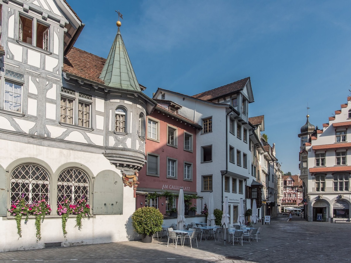 A charming square is depicted, featuring a collection of traditional buildings with distinctive architecture. Cafes and tables are visible in the foreground, surrounded by flower arrangements. The scene is bathed in natural light, with a clear blue sky above.