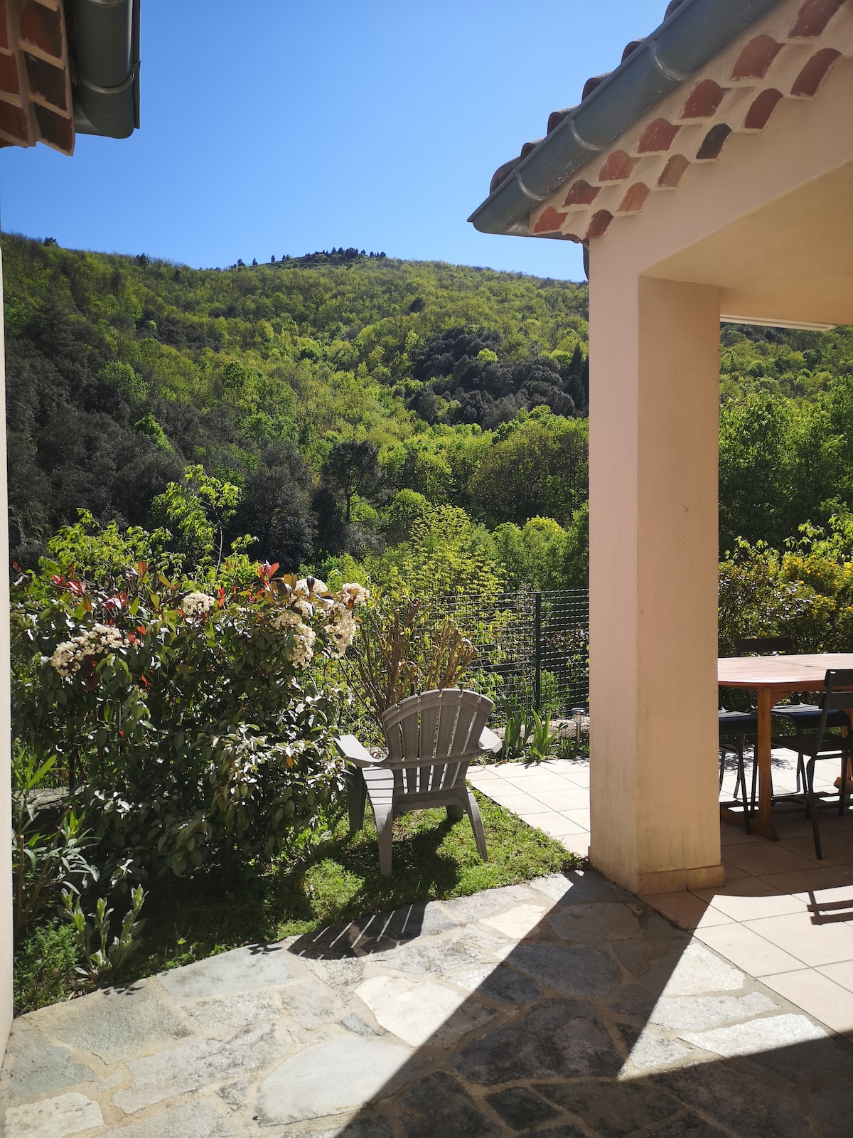 A serene outdoor space is seen with a curved stone pathway leading to a covered terrace. A wooden table is set under a canopy, and a gray lounge chair is positioned amid lush greenery. Rolling hills are visible in the background under a clear blue sky.