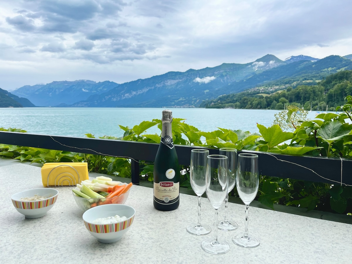 A table is set on a balcony overlooking a tranquil lake, with mountains in the background. Four champagne flutes are arranged beside a bottle of sparkling wine. Bowls of vegetables and a creamy dip are positioned on the table, surrounded by vibrant greenery.