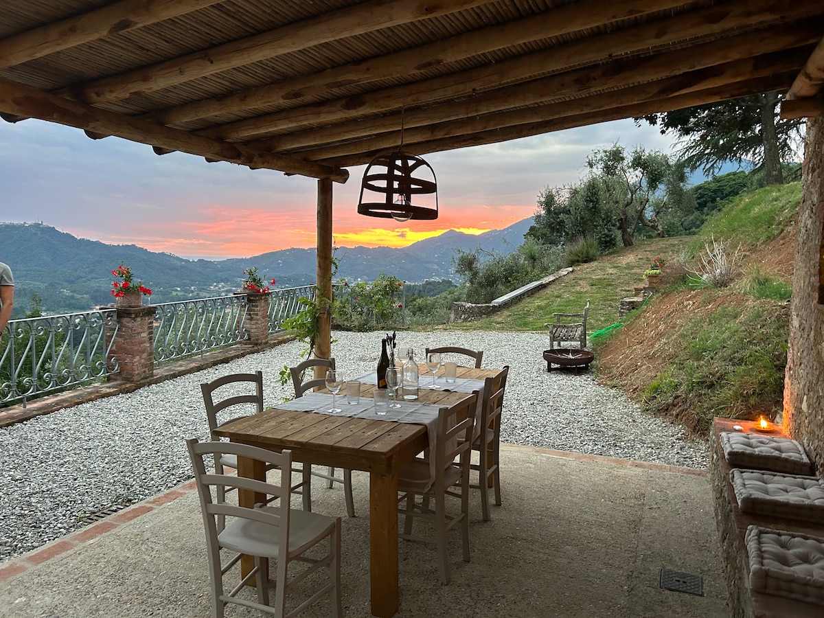 An outdoor dining area is presented under a rustic wooden pergola, featuring a large table surrounded by white chairs. Scenic views of rolling hills are visible in the background, with vibrant colors from the sunset illuminating the sky.