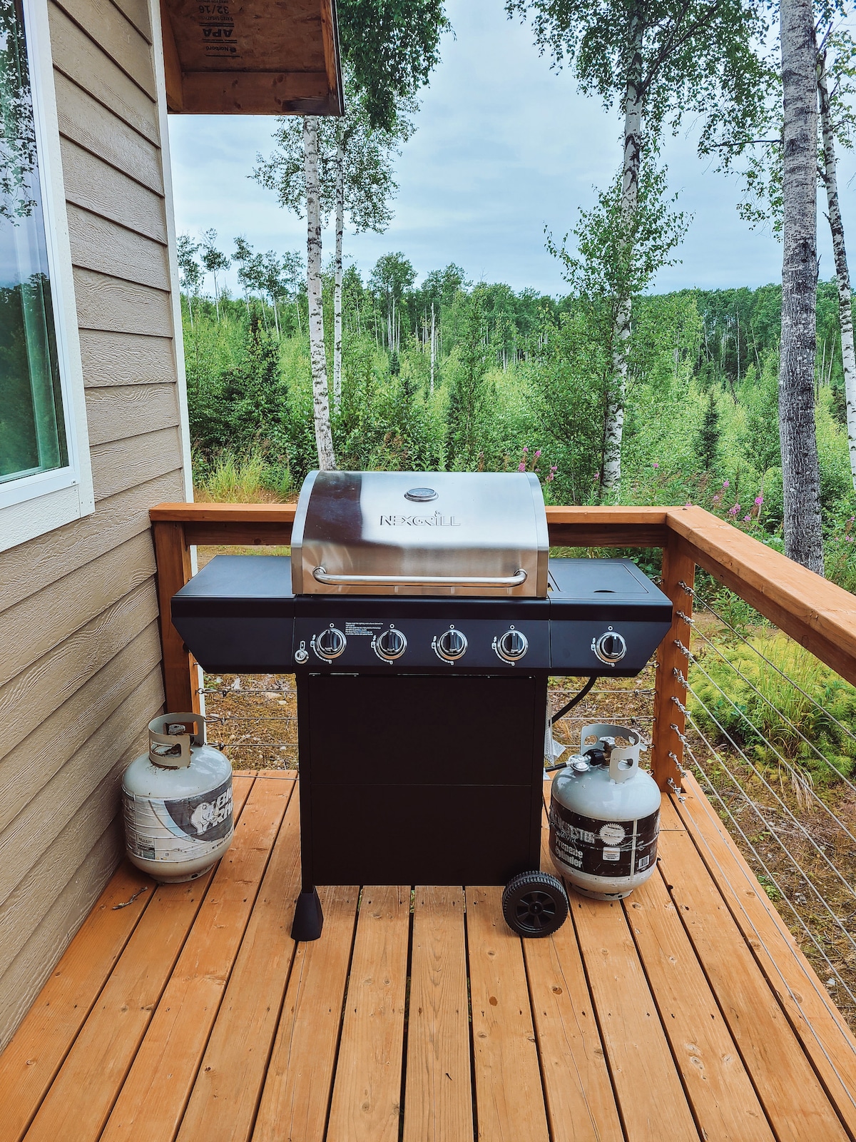 A gas grill is situated on a wooden deck, flanked by two propane tanks. Surrounding the deck, lush greenery and trees are present, creating a natural backdrop. The sky above is overcast, adding to the serene outdoor atmosphere.