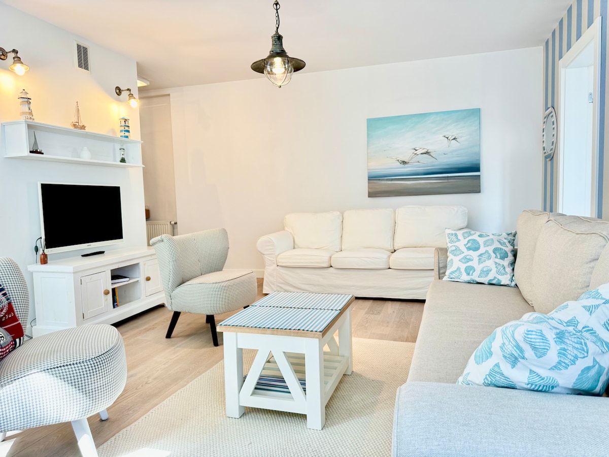 A modern living room is depicted with two comfortable sofas, a light-colored armchair, and a coffee table. Natural light enters through spacious windows, highlighting the soft color palette. A television is positioned on a white console, and art featuring seagulls decorates the wall.