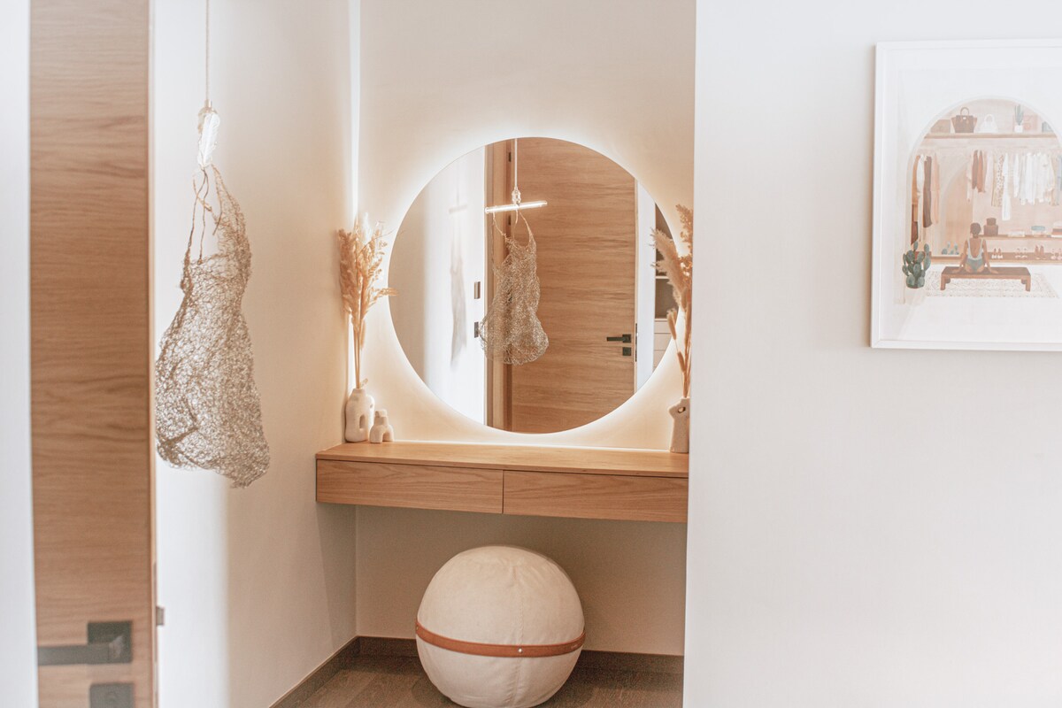 A well-lit entryway features a circular mirror with integrated lighting, reflecting warmth. A wooden console table is positioned beneath the mirror, adorned with decorative elements. A plush round pouf sits invitingly on the floor, adding a touch of comfort to the welcoming space.