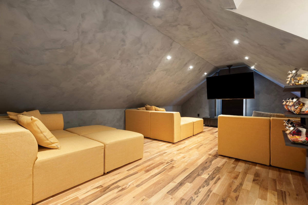 An inviting loft space features two spacious yellow couches arranged for relaxation and socializing. The warm wood flooring complements the modern design, while recessed lighting brightens the room. A large screen television is mounted on the opposite wall, enhancing the entertainment atmosphere.