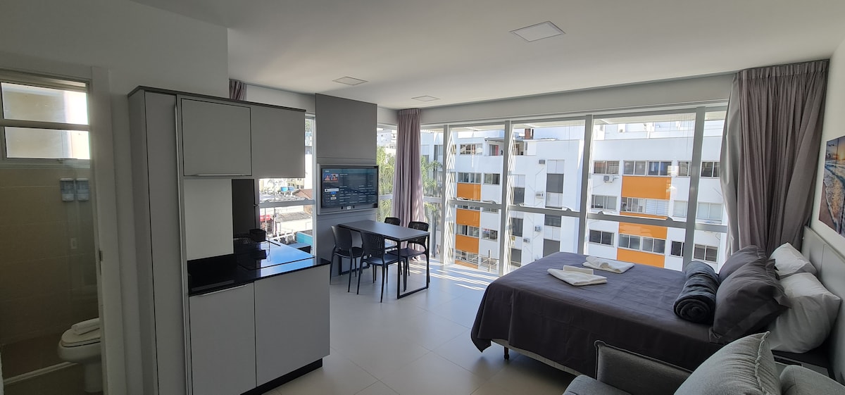 A bright studio apartment features large windows allowing natural light to fill the space. An open kitchen area is complemented by a dining table and chairs. A spacious bed is arranged with folded towels, while a television is mounted on the wall across from the seating area.