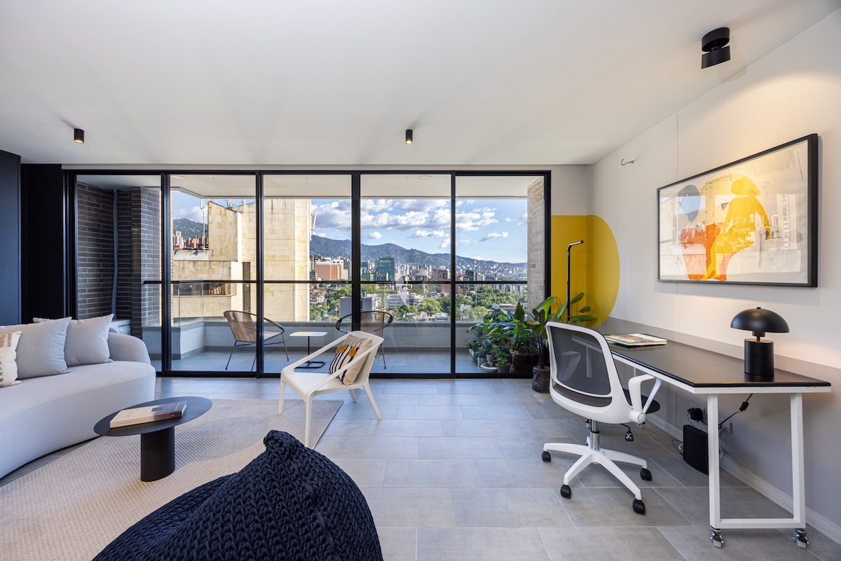 An inviting living area features a modern design with large glass doors opening to panoramic views of Medellín. A spacious seating area includes a light-colored couch and a black accent table. A workspace with a white desk and ergonomic chair is set beside lush greenery, enhancing the ambiance.