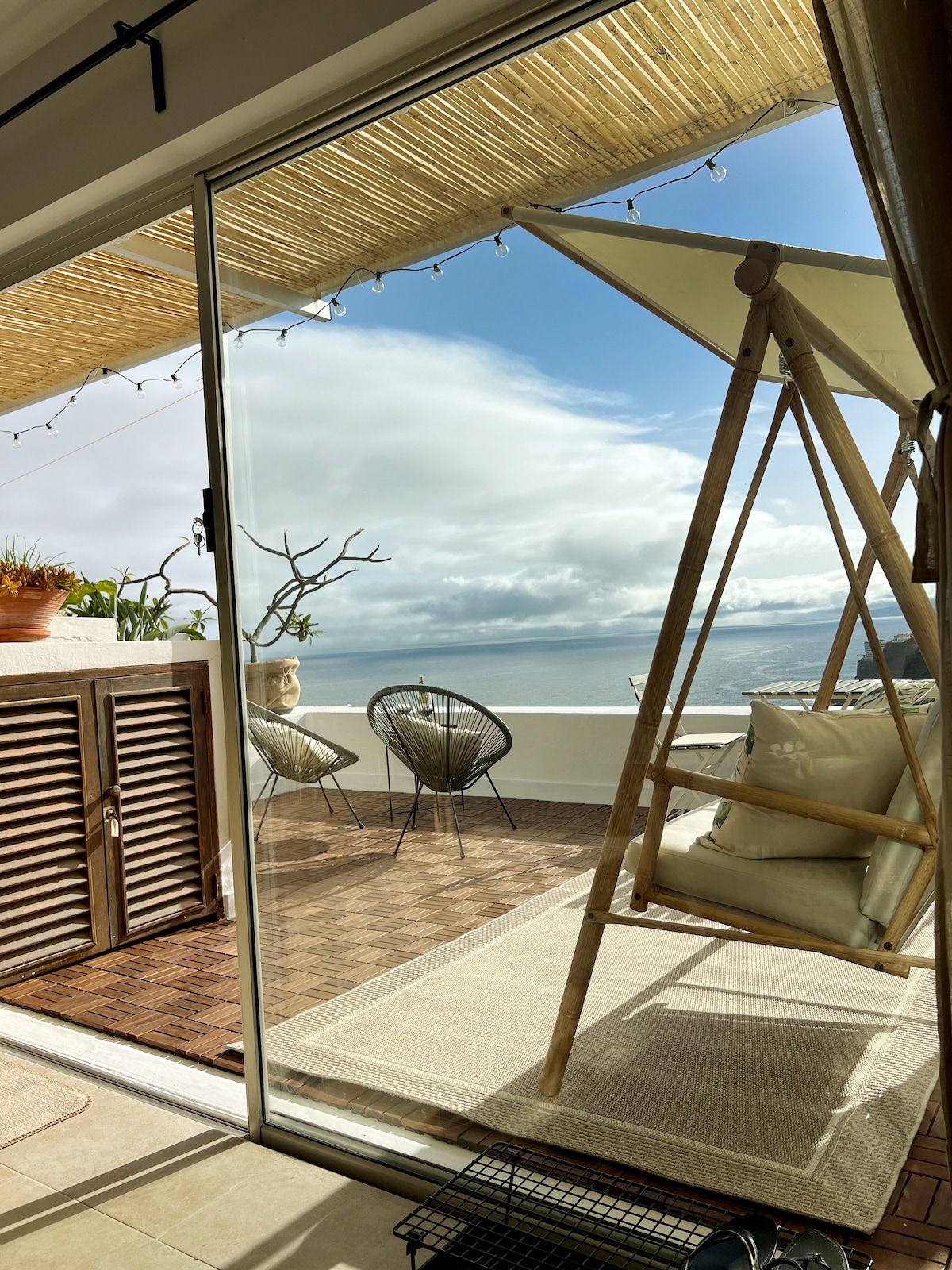 A light-filled space reveals a private terrace with a swing chair and stylish seating. The terrace features a calming view of the ocean and is adorned with greenery. Large glass doors allow for seamless indoor-outdoor flow, enhancing the tranquil atmosphere.
