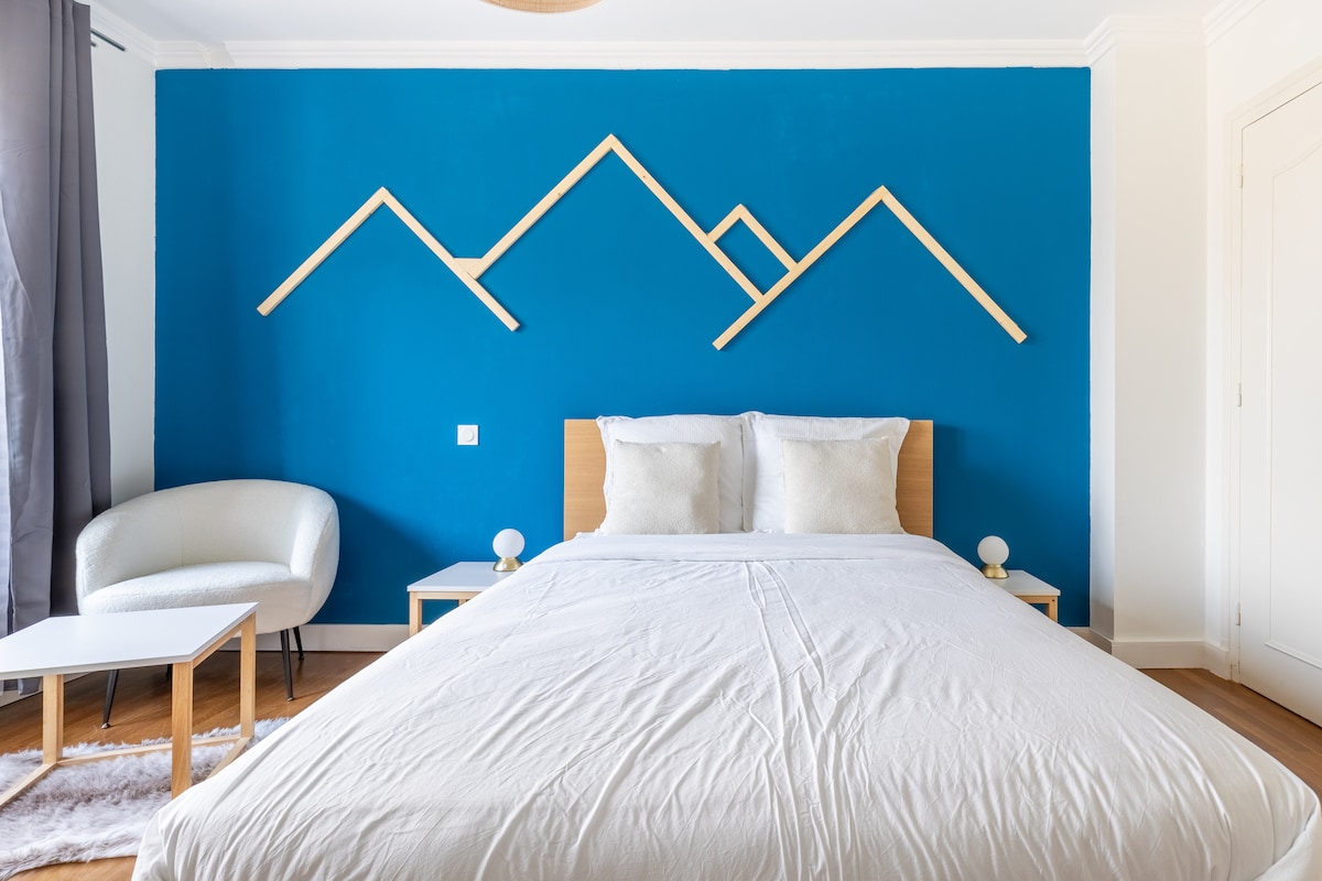 A spacious bedroom features a light-colored bed adorned with white linens, complemented by two white cushions. An accent wall in a vibrant blue is decorated with geometric wooden shapes, while a cozy armchair and small tables are arranged nearby.