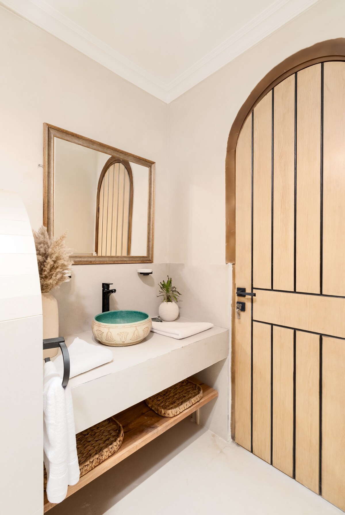 The bathroom features a minimalist design with a natural stone basin atop a smooth countertop. A large mirror with an arched frame reflects the soft colors of the room. Simple wooden shelves hold woven baskets and fresh towels, enhancing the tranquil ambiance.
