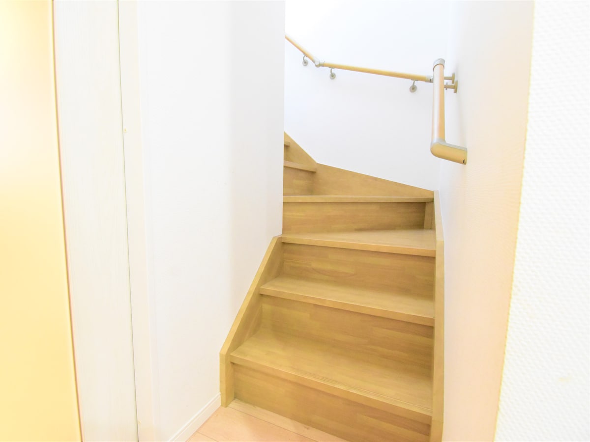 A wooden staircase is depicted, featuring smooth steps and a light-coloured handrail. The walls are white, enhancing the bright and open appearance of the space. Natural light is suggested to be present, contributing to a welcoming atmosphere.