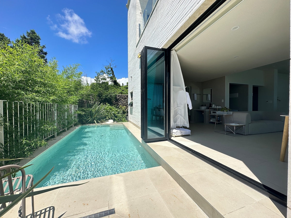 An outdoor area showcases a private pool with crystal-clear water, bordered by light stone tiles. Large glass doors open to an inviting interior space, allowing natural light to fill the area. Lush greenery surrounds the pool, contributing to a serene atmosphere.