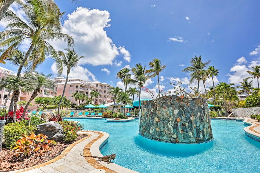 A tropical resort pool features a stone centerpiece surrounded by clear blue water. Lush palm trees and vibrant landscaping create a relaxing atmosphere. Loungers and umbrellas are positioned around the pool, offering shaded seating options in a sunlit environment.