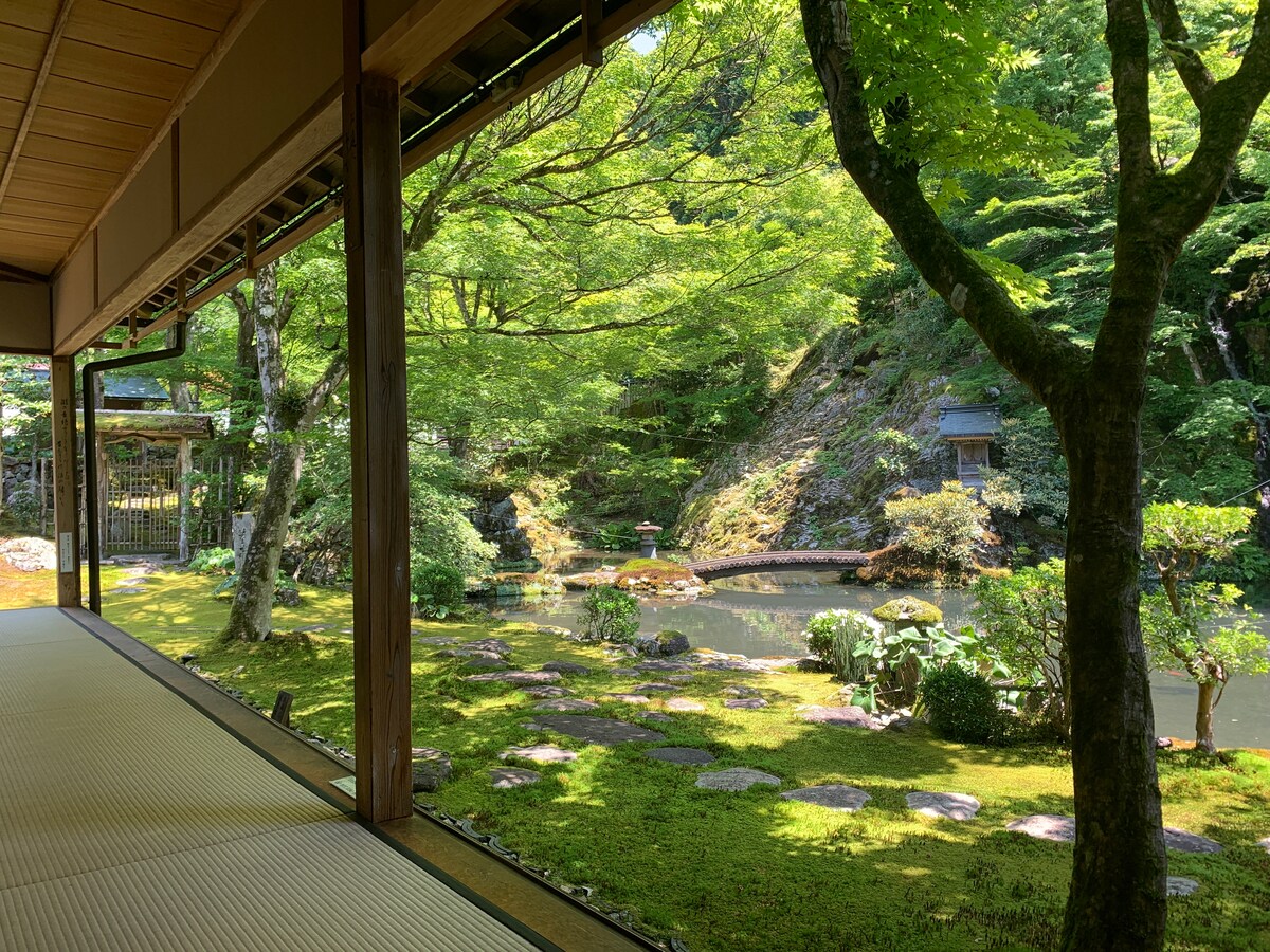 A serene view from the interior of the traditional house showcases a lush garden with vibrant greenery surrounding a tranquil pond. The sunlight filters through the trees, creating a peaceful atmosphere. Stone pathways meander through the garden, leading to various landscape features.