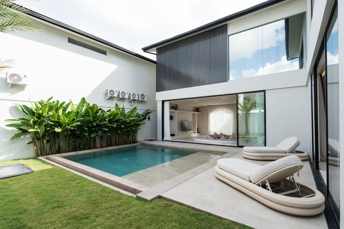 The outdoor area features a private pool surrounded by neatly manicured grass. Sun loungers are positioned beside the pool, offering a space to relax. The villa's modern design is evident in the large glass windows and sleek architecture, complemented by lush green plants in the background.