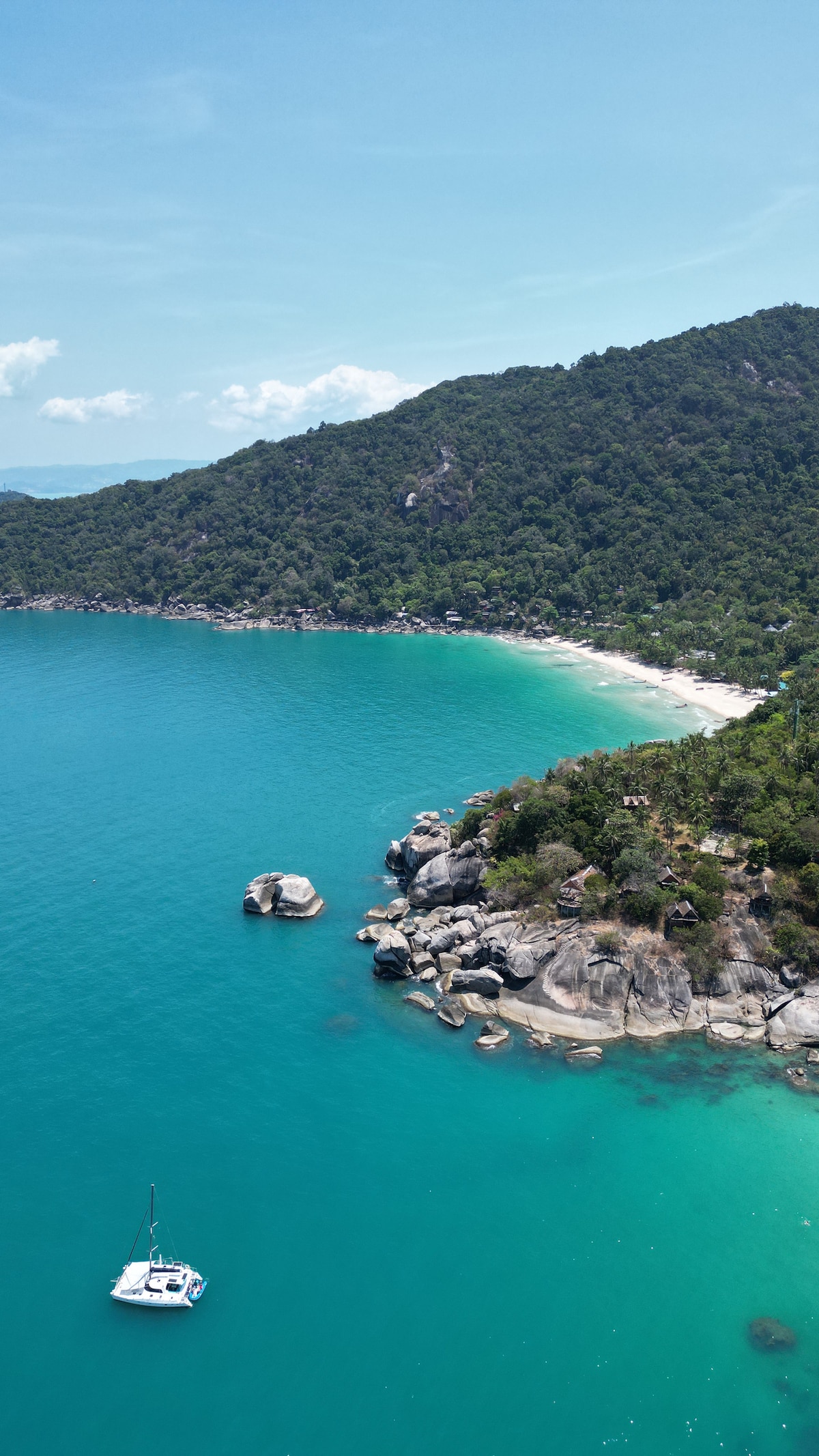 A panoramic view reveals a secluded beach lined with lush greenery and rocky formations. Clear turquoise waters gently lap at the shore, where a small sailboat is anchored. The landscape showcases a harmonious blend of natural beauty and tranquility.