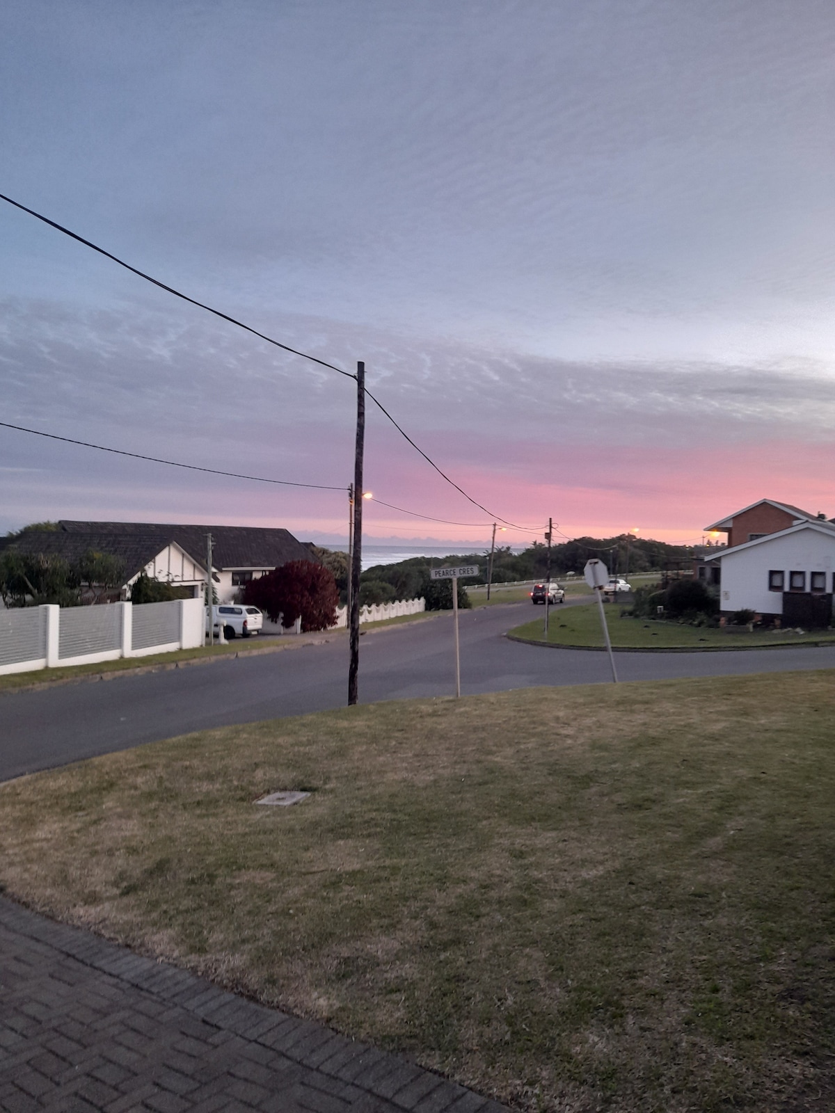 A serene street view captures a gentle gradient of colors in the sky during twilight, with soft purples and pinks blending together. Houses are seen along the road, alongside grassy areas and a few scattered trees, creating a tranquil neighborhood setting.