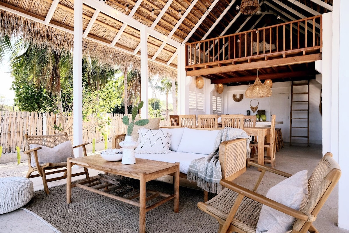 An open-air living area features natural materials, with a thatched roof and wooden beams creating a relaxed atmosphere. A comfortable seating arrangement includes a spacious sofa, two woven chairs, and a coffee table. The dining area is visible in the background, complemented by handcrafted lighting.
