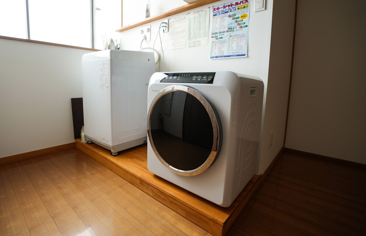 A modern laundry area is showcased with a sleek washing machine featuring a large, circular door. Next to it, a compact refrigerator is positioned against the wall. Natural light streams through a nearby window, highlighting the clean wooden flooring.