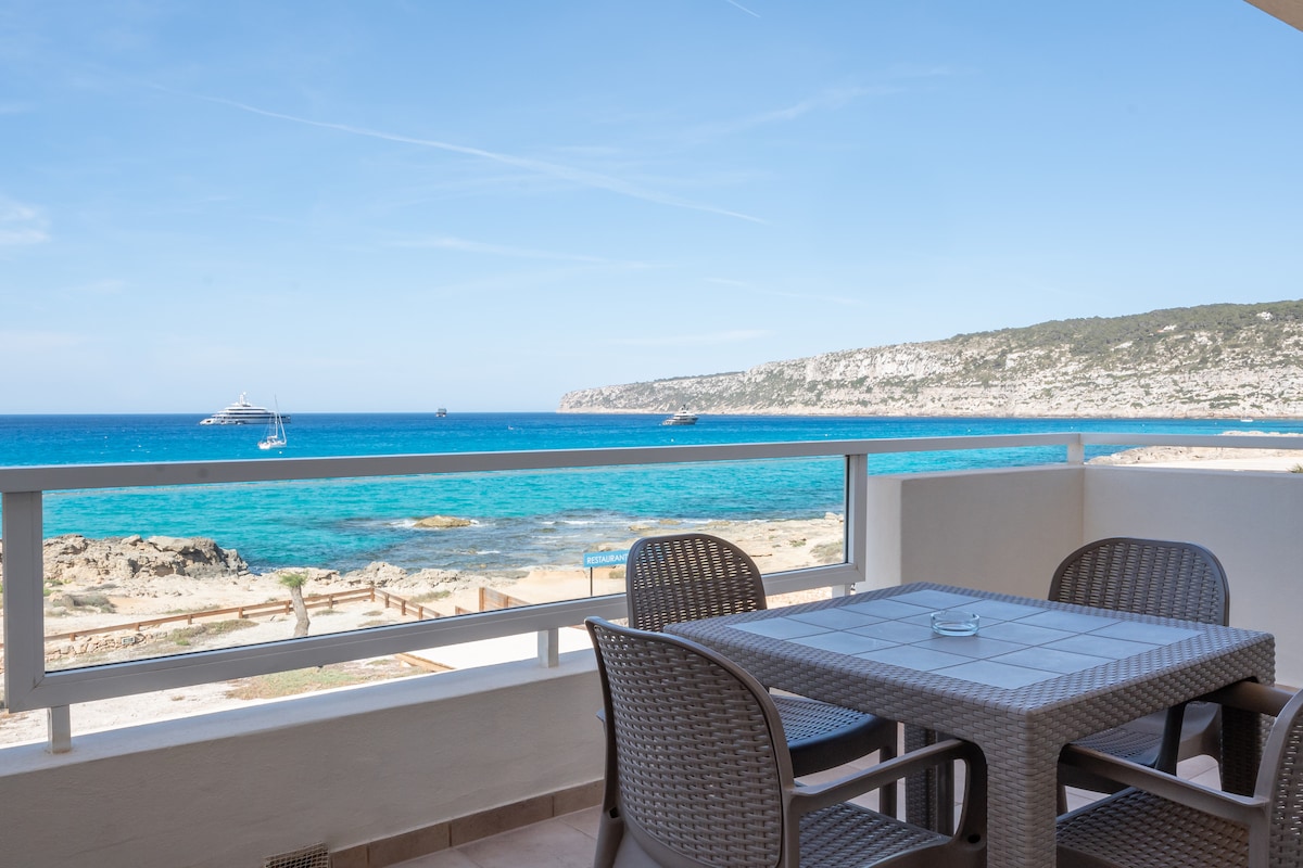 A balcony is depicted, featuring a modern table surrounded by four woven chairs. The turquoise sea stretches into the distance, with a clear sky above. Sailboats can be seen gently floating on the water, and rocky hills rise in the background.