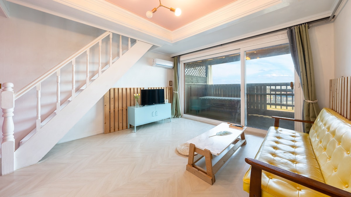 A bright living area features a combination of a yellow sofa and a wooden coffee table. Large sliding glass doors provide ample natural light and open to a balcony with views. A staircase leads to the upper level, while light wood paneling adds a subtle touch.