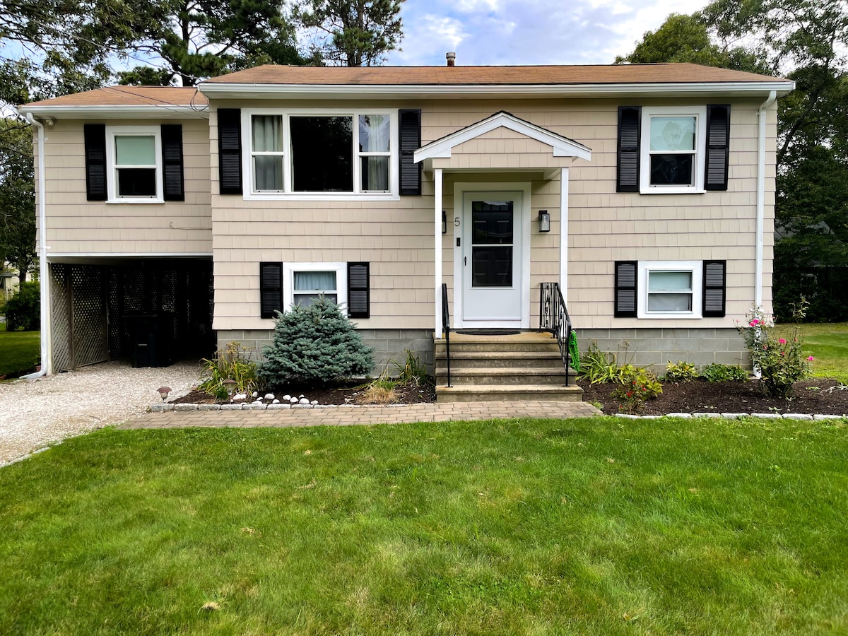 The exterior of the home is featured, showcasing a two-story design with light-colored siding and dark shutters. A welcoming front porch is visible, along with a neatly landscaped yard that includes shrubs and flowers. The driveway is gravel, and a pathway of bricks leads to the entrance.