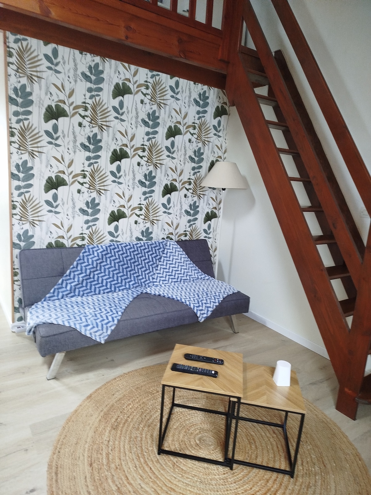 A cozy seating area is presented with a gray sofa adorned with a blue throw blanket. A small wooden table sits in front, accompanied by a round rug. A standing lamp provides light, and a wooden staircase leads to an upper level, enhancing the vertical space.