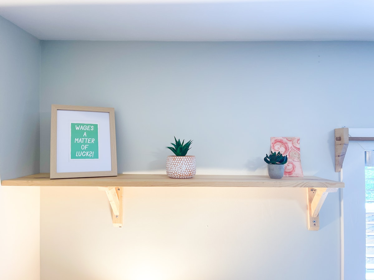 A wooden shelf is mounted on a light-colored wall, displaying a framed print with motivational text, a textured planter with a green succulent, and a small decorative piece. Additional greenery is present in another small planter, contributing to the serene decor.