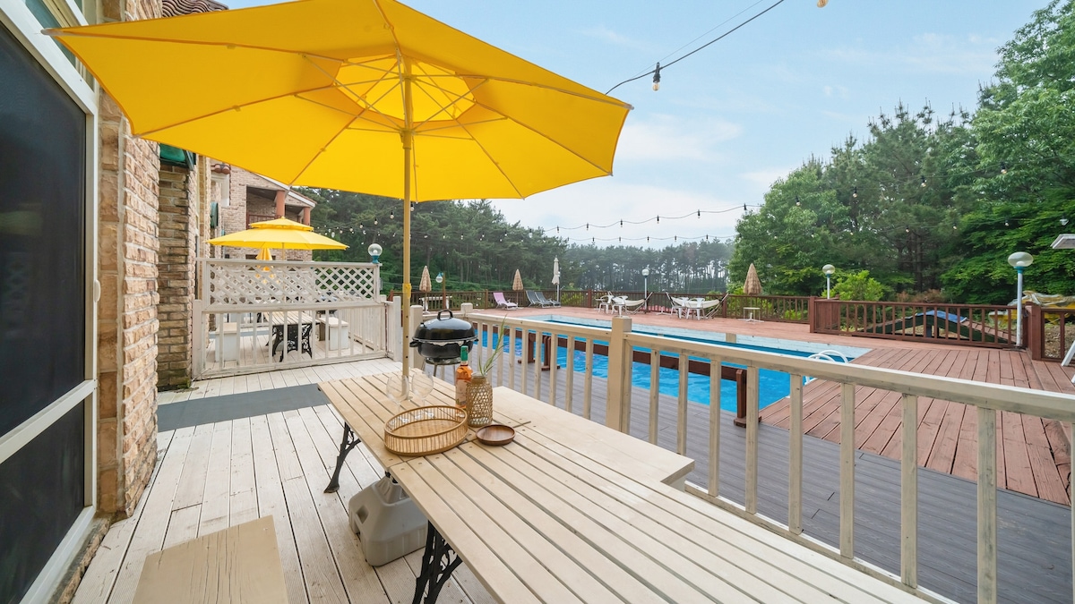 A spacious outdoor deck features a long wooden table shaded by a large yellow umbrella. A grill is positioned nearby, with a private pool visible in the background. Lush greenery and additional seating areas enhance the open atmosphere.