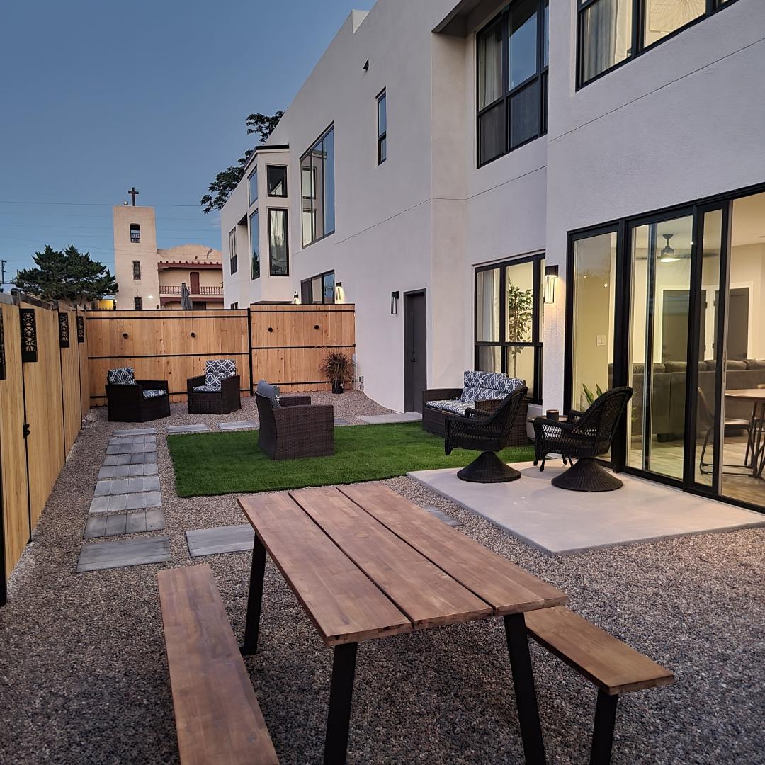 An outdoor space showcases a combination of seating options, including wicker chairs and a picnic table. Soft grass patches are bordered by a stone walkway, while warm lighting enhances the ambiance during evening hours.