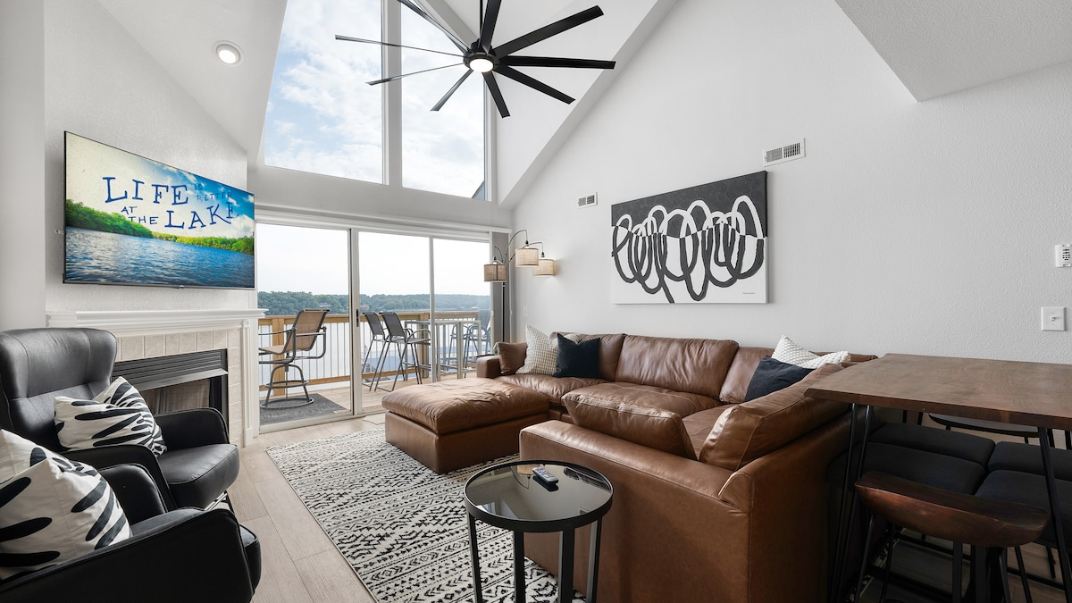 A spacious living area features a large sectional sofa facing a flat-screen TV mounted above a fireplace. Large windows allow natural light to fill the space, showcasing a stunning view of the lake. A dining table and chairs are positioned nearby, enhancing the room's functionality.