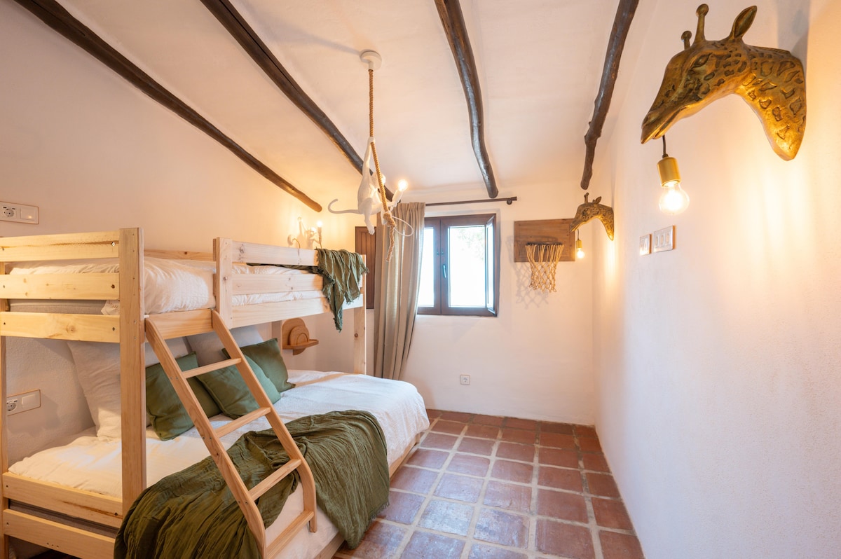 Dormitorio con litera de madera para tres personas, colchones con sábanas y mantas verdes, vigas oscuras en techo, lámparas colgantes, ventana con cortinas, suelo de baldosas rústicas y aro de baloncesto decorativo en la pared.
