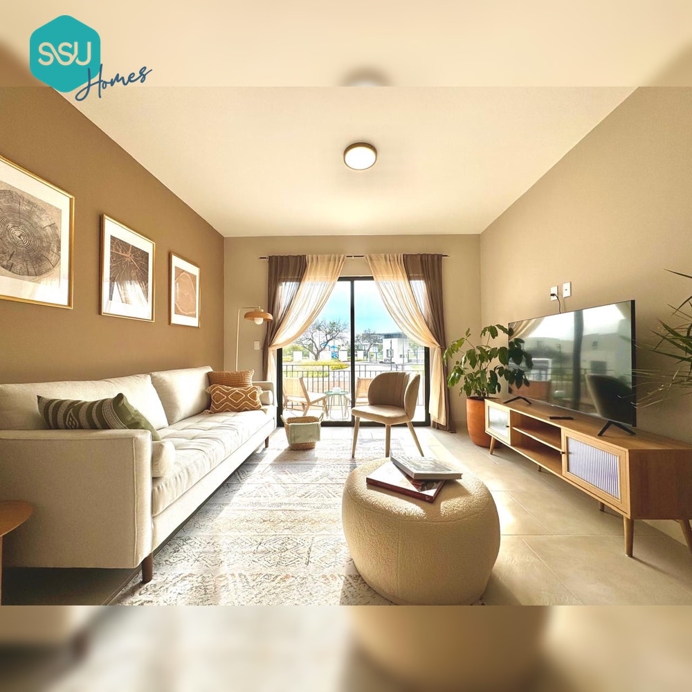 A comfortable living area is furnished with a light-colored sofa and a wooden coffee table. Large glass doors allow natural light to fill the space and provide access to a balcony. A television is positioned on a modern media console, complemented by decorative wall art.