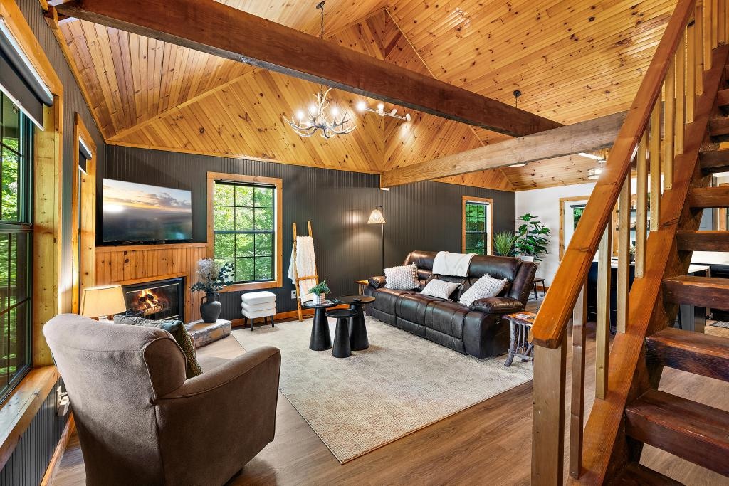 A cozy living room features natural wood accents and high ceilings. A large couch and armchair invite relaxation, positioned around a gas fireplace. Large windows frame views of the outside greenery, while soft lighting enhances the welcoming ambiance of the space.