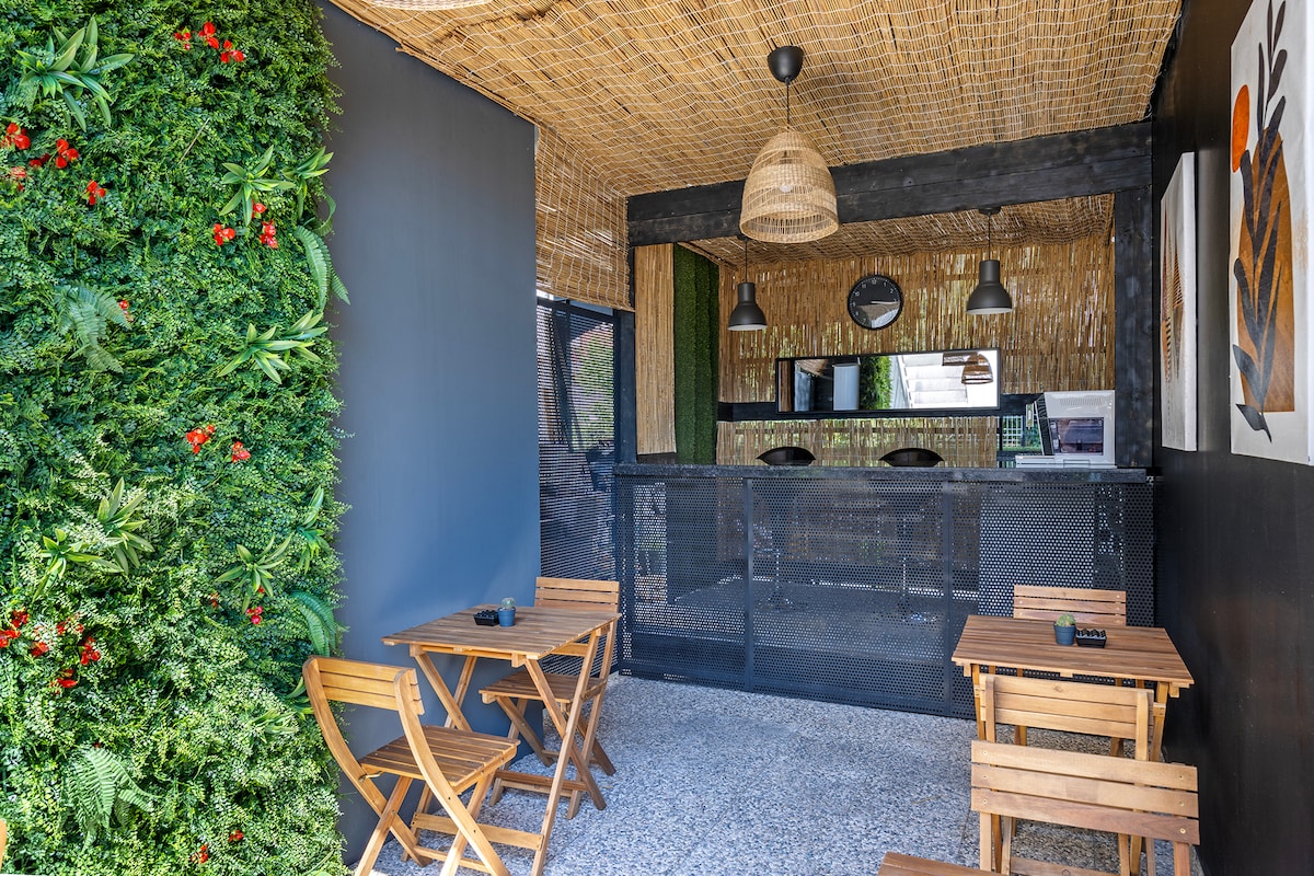 A casual outdoor seating area is depicted, featuring two wooden tables and matching chairs. A green wall adorned with vibrant foliage adds a natural touch, while a bamboo roof provides shade. A sleek service area is visible in the background with a round mirror reflecting light.