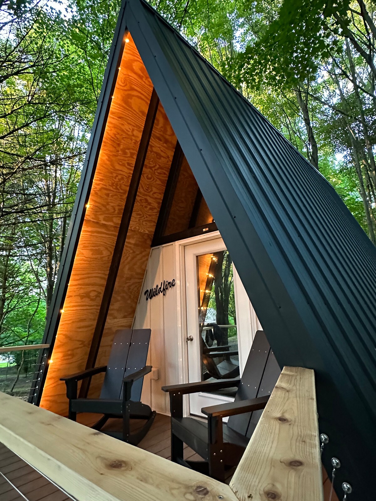 An A-frame cabin stands amidst lush greenery, featuring a distinctive triangular roof. Two wooden chairs are positioned on the porch, inviting relaxation. Warm lighting highlights the entrance, while a sign labeled 'Wildflower' adds a personal touch to the cabin's exterior.