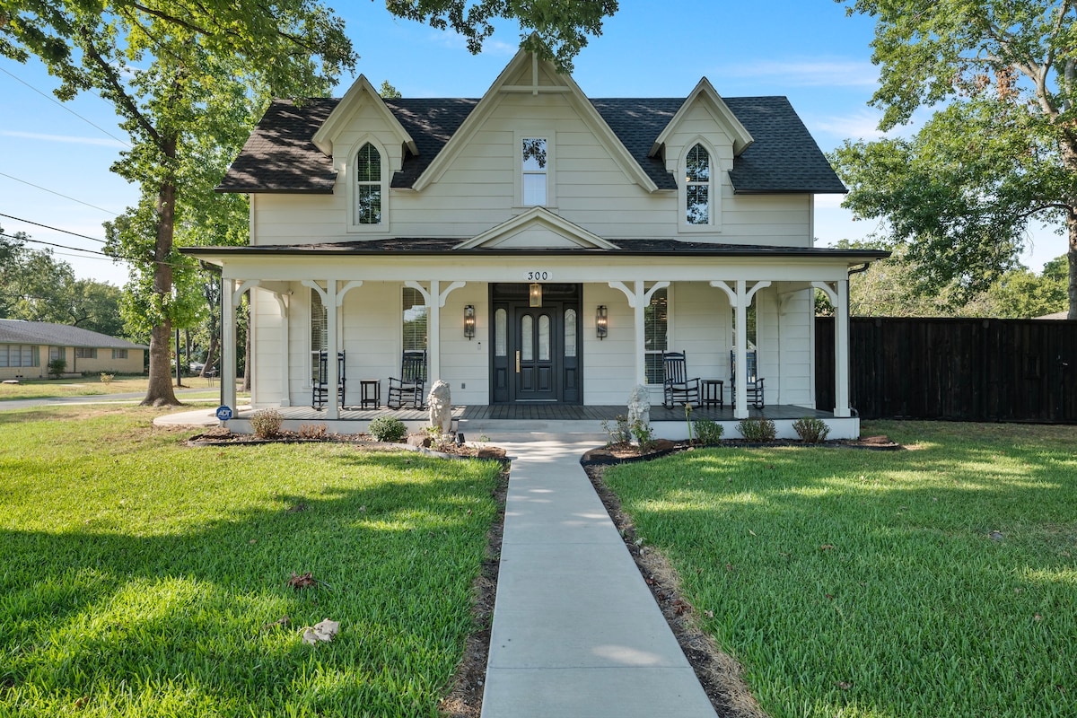 A charming Victorian home is set amidst lush green lawn and trees. The front porch, furnished with rocking chairs, invites relaxation. A paved walkway leads to the entrance, framed by floral landscaping, enhancing the inviting exterior of the house.