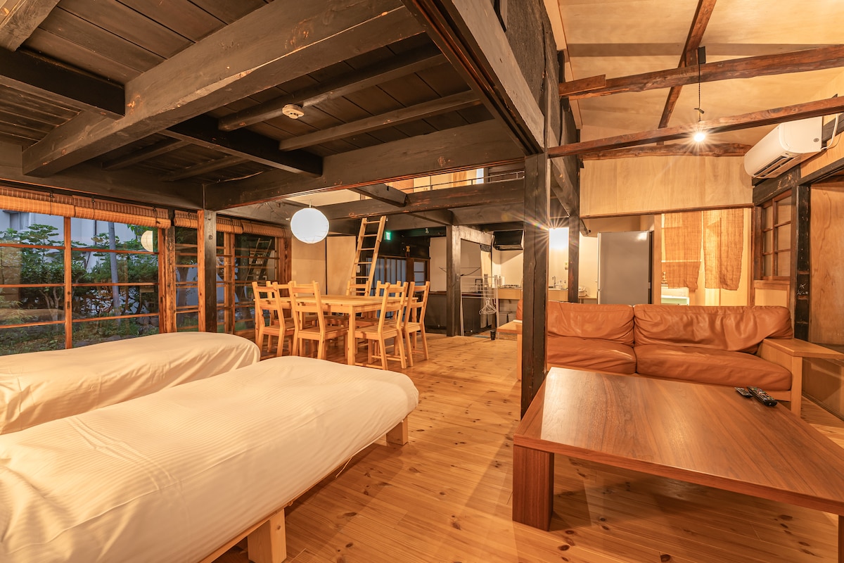 An open living space features two spacious single beds and a comfortable leather sofa. Natural wood tones dominate the interior, with a dining table set for six. Large windows allow sunlight to brighten the room, enhancing the connection to the outdoors.