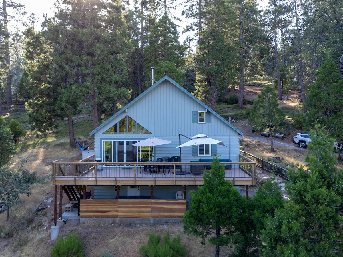 The cabin is surrounded by tall pine trees and features a spacious deck with outdoor seating. Large sliding glass doors allow natural light to brighten the interior, showcasing a modern design. The area displays a mix of greenery and landscaped outdoor space.