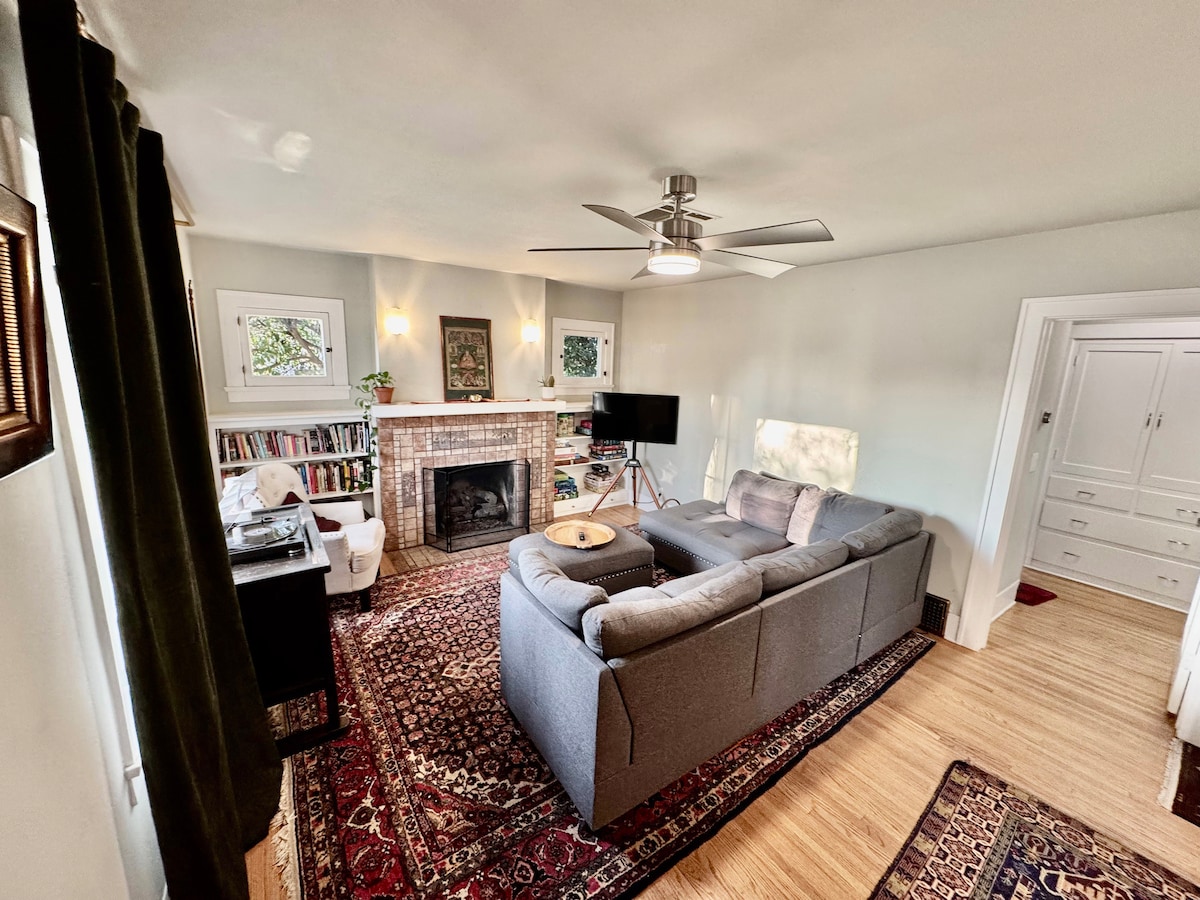 A living room features a comfortable gray sofa facing a gas fireplace set within a tiled mantel. Light wooden flooring complements the area, and a ceiling fan is visible above. Shelving filled with books and a television stand are situated nearby, enhancing the cozy environment.