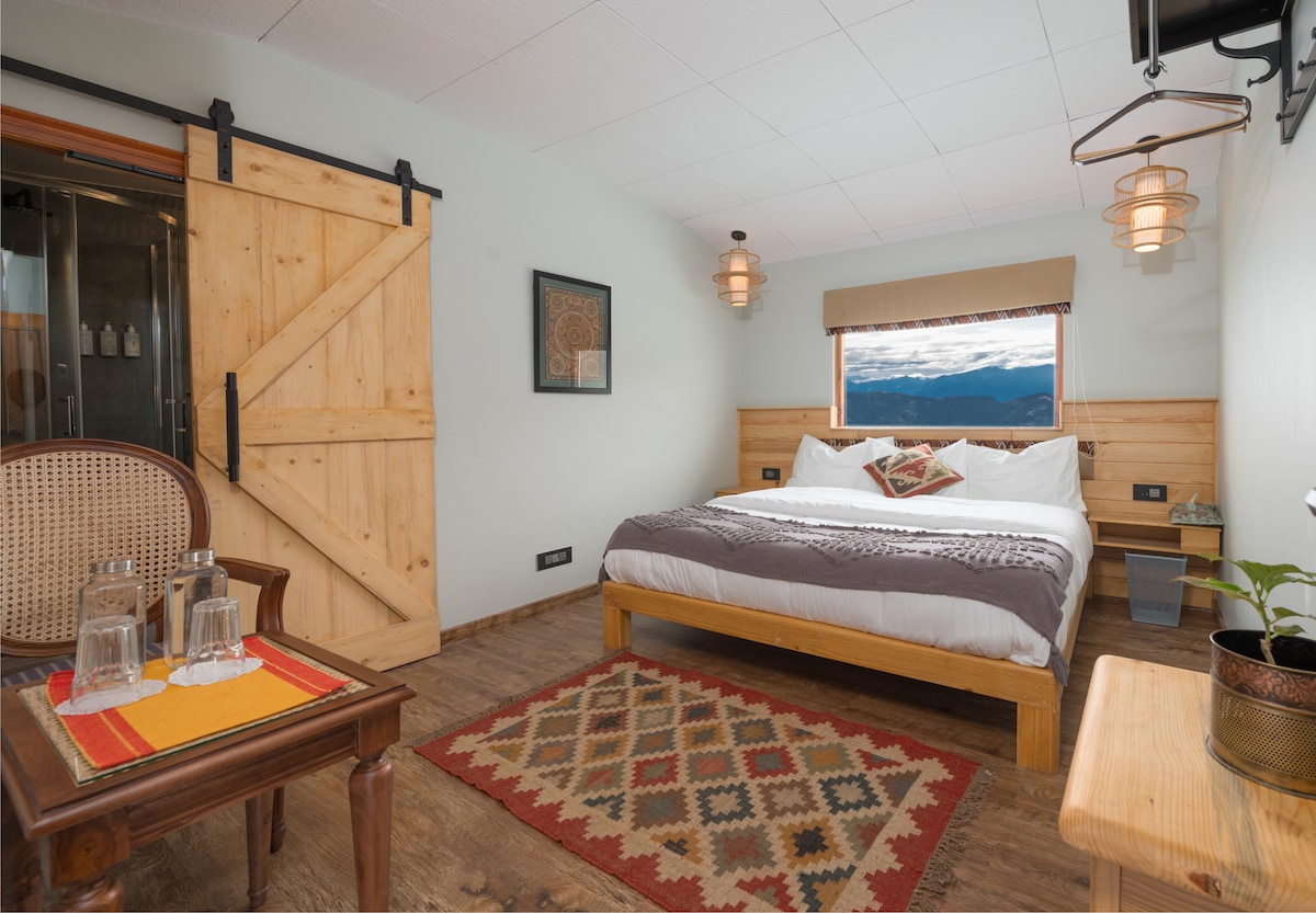 A cozy room features a spacious bed with crisp white linens and decorative pillows. Warm wooden accents and rustic decor are highlighted by ambient lighting. A patterned rug lies beneath a polished wooden floor, and a small table with water glasses is positioned nearby.