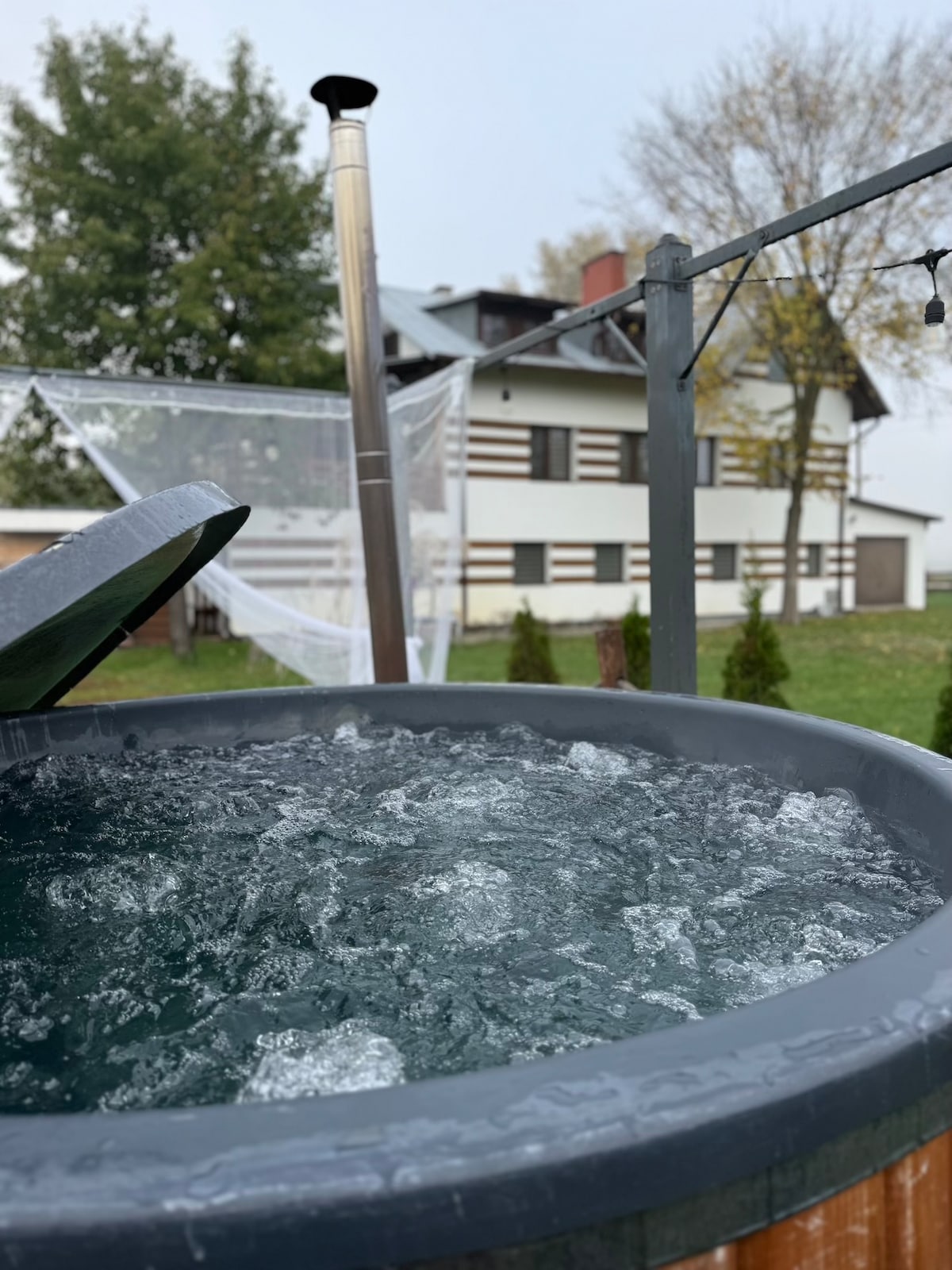 A wooden hot tub is filled with bubbling water, positioned in the foreground. In the background, a multi-story house is visible, featuring a mix of light and dark wooden siding, surrounded by a green lawn and small trees.