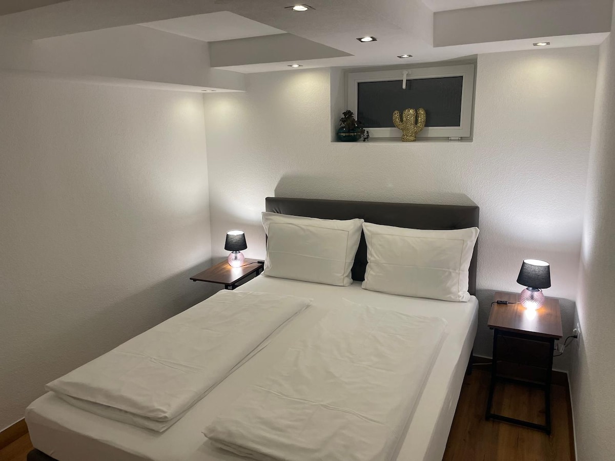 A cozy sleeping area is depicted, featuring a spacious bed with white linens and two neatly placed towels. On either side of the bed, small tables with lamps provide soft illumination. A window is visible above the headboard, contributing to the overall brightness of the room.