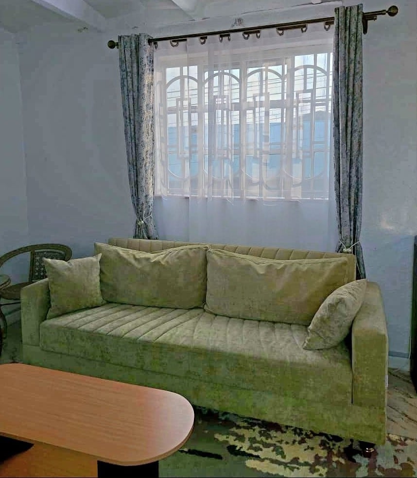 A soft green sofa is positioned in a cozy corner, complemented by decorative pillows. Natural light filters through a window adorned with patterned curtains, highlighting the neutral tones of the surrounding decor.