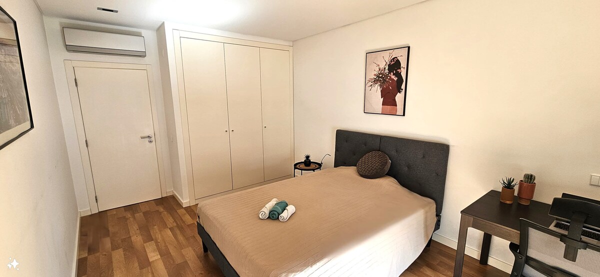 A well-organized bedroom is presented, featuring a spacious bed with a neutral-toned bedspread. A piece of artwork adorns the wall above the bed. A small table with decorative plants sits beside the bed, and a work desk is positioned near a window.