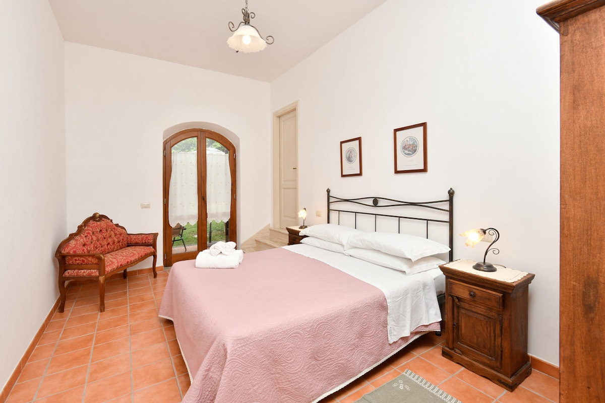 Тази уютна спалня разполага с удобно двойно легло с меко розово спално бельо, подова настилка от теракотени плочки и очарователен декор. Френските врати се отварят към живописна гледка, а диван във венециански стил, нощна лампа, климатик и гардероб добавят комфорт.