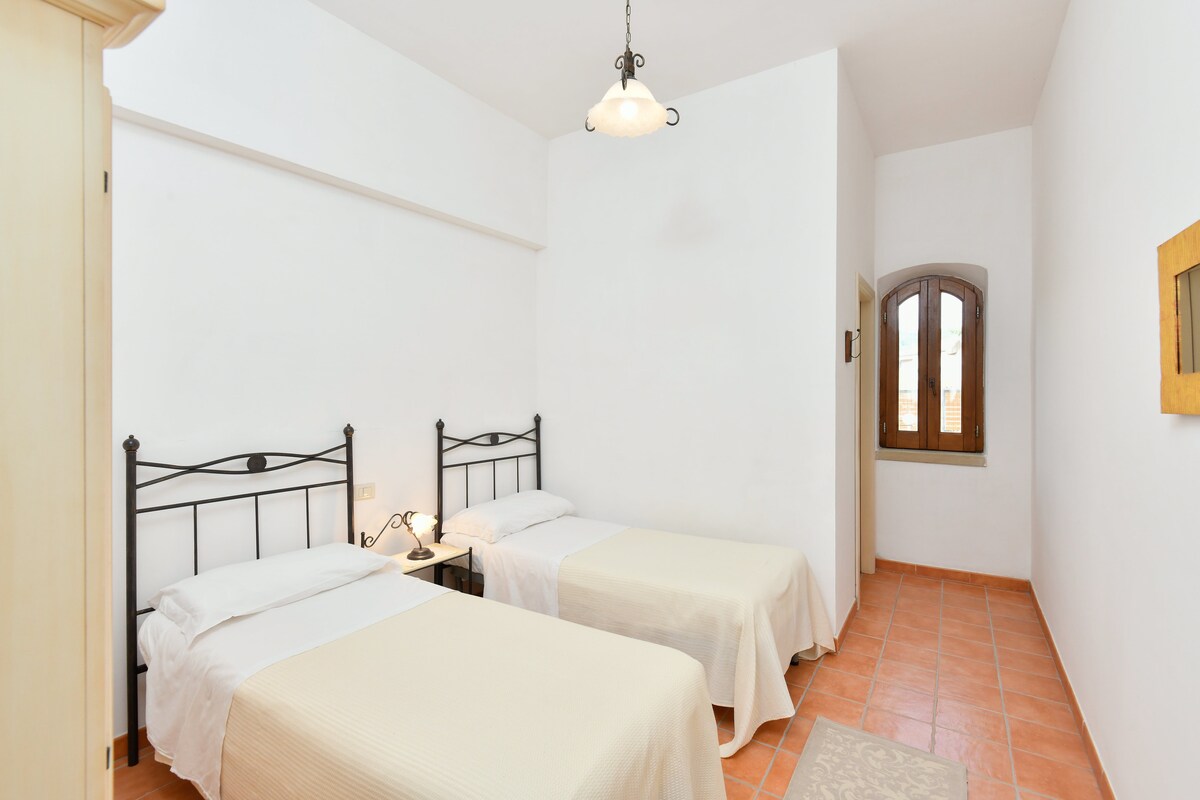 Тази уютна спалня разполага с две единични легла с меко бежово спално бельо, подова настилка от теракотени плочки, климатик и гардероб. Очарователна нощна лампа и малък прозорец внасят естествена светлина, създавайки спокойна атмосфера за релаксиращ престой.