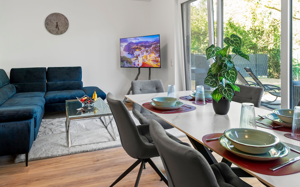 An inviting dining area and living space are featured, with a modern glass table set for four. Comfortable chairs surround the table, and a blue sectional sofa is positioned nearby. Large windows allow natural light to fill the room, enhancing the overall ambiance.