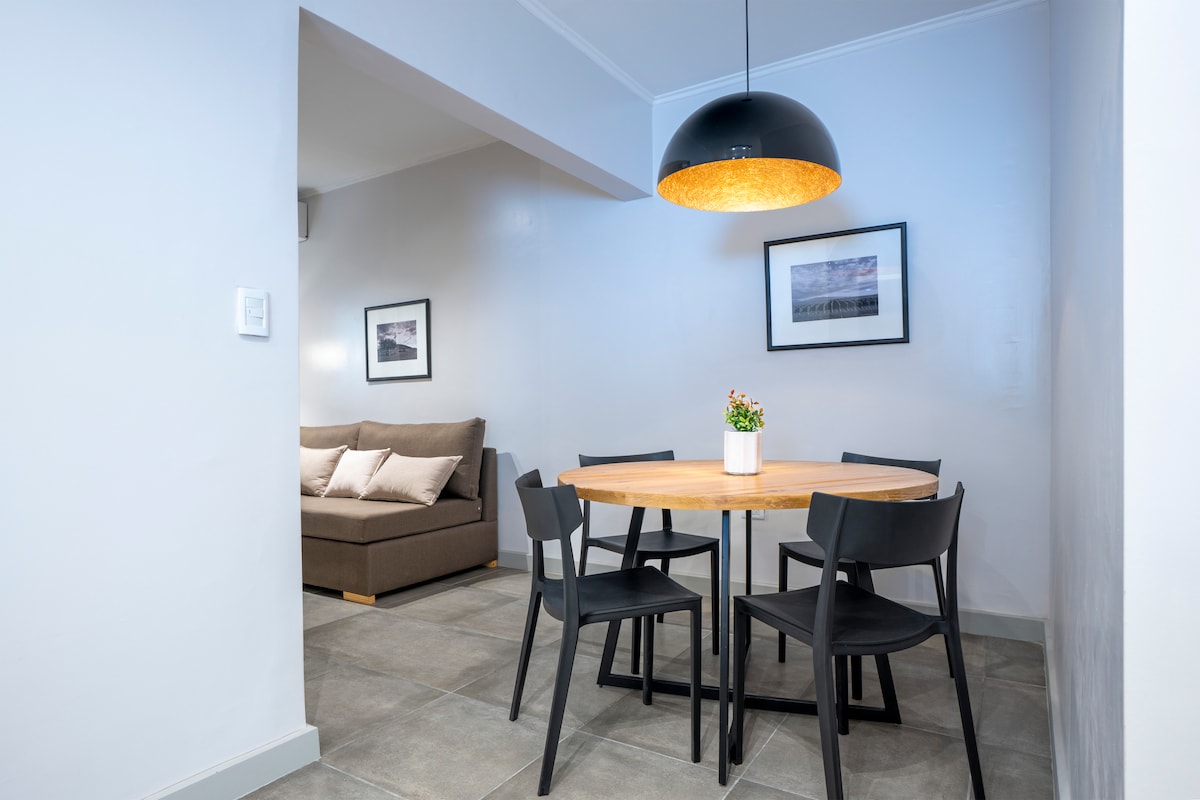 A dining area is presented with a round wooden table surrounded by four black chairs. A soft, neutral-toned sofa is positioned nearby. Above, a modern pendant light hangs, and two framed photographs adorn the walls, adding subtle decor to the space.